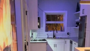 Cocina compacta con gabinetes blancos, estufa a gas, lavamanos y una ventana con cortinas.