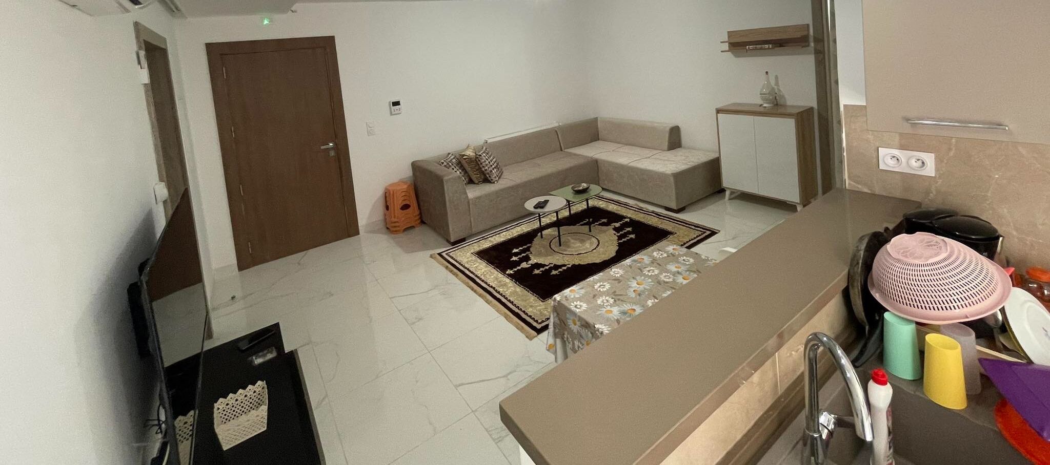 A modern living room with a sofa, a coffee table, and a rug. There is a door, a small cabinet, and a countertop with kitchen items.