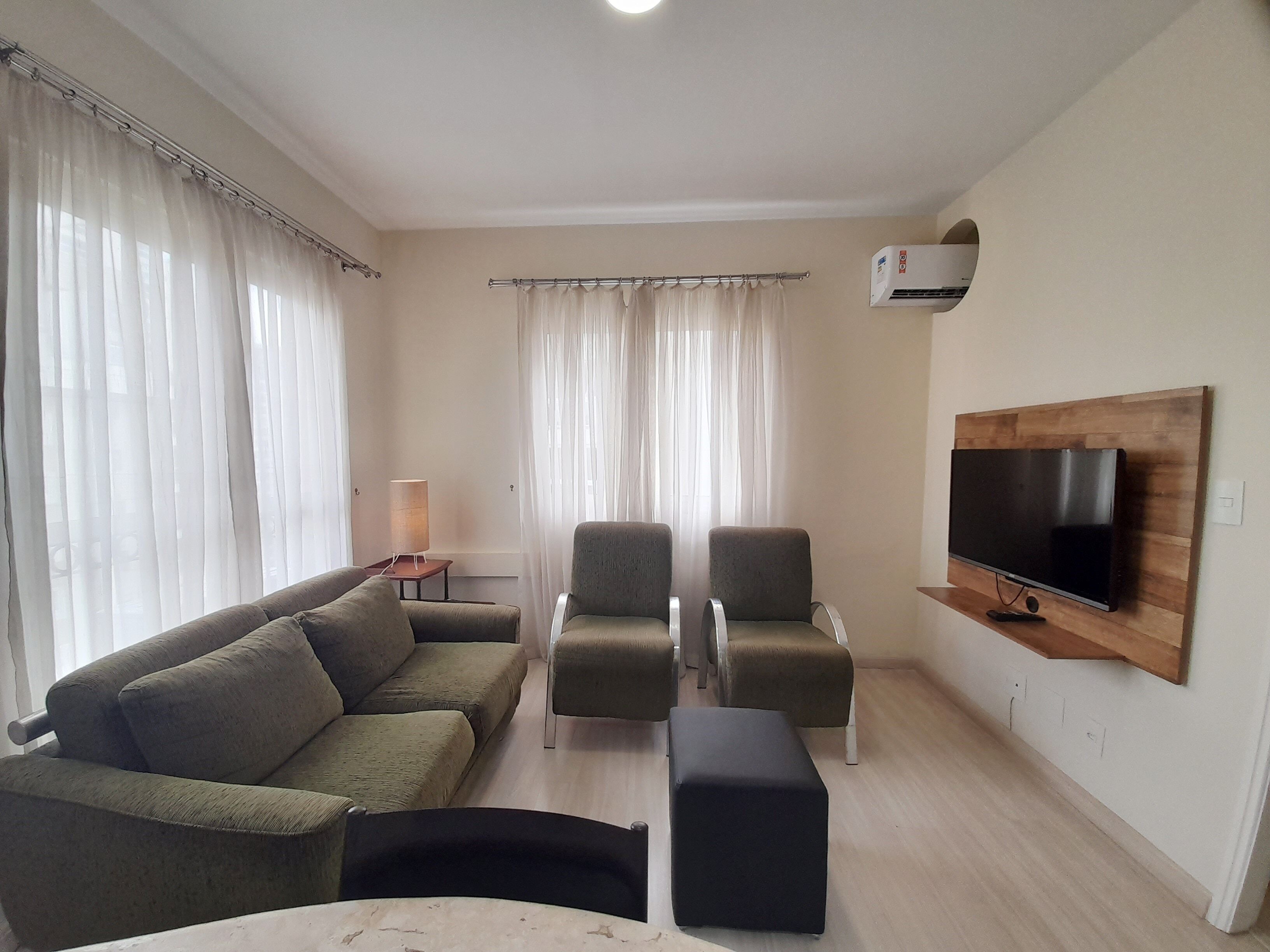 Un salon comprenant un canapé, deux fauteuils, une table basse, un téléviseur fixé sur un panneau en bois et une lampe sur une table d’appoint.