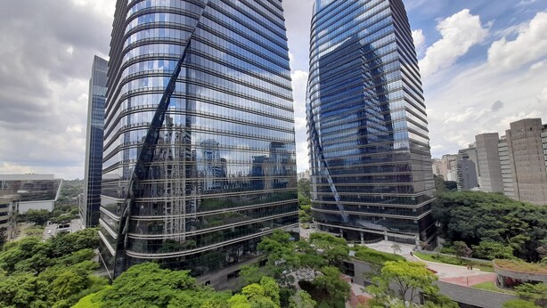 Dos edificios modernos de gran altura con fachadas de vidrio reflectante, rodeados de exuberante vegetación y otras construcciones urbanas.