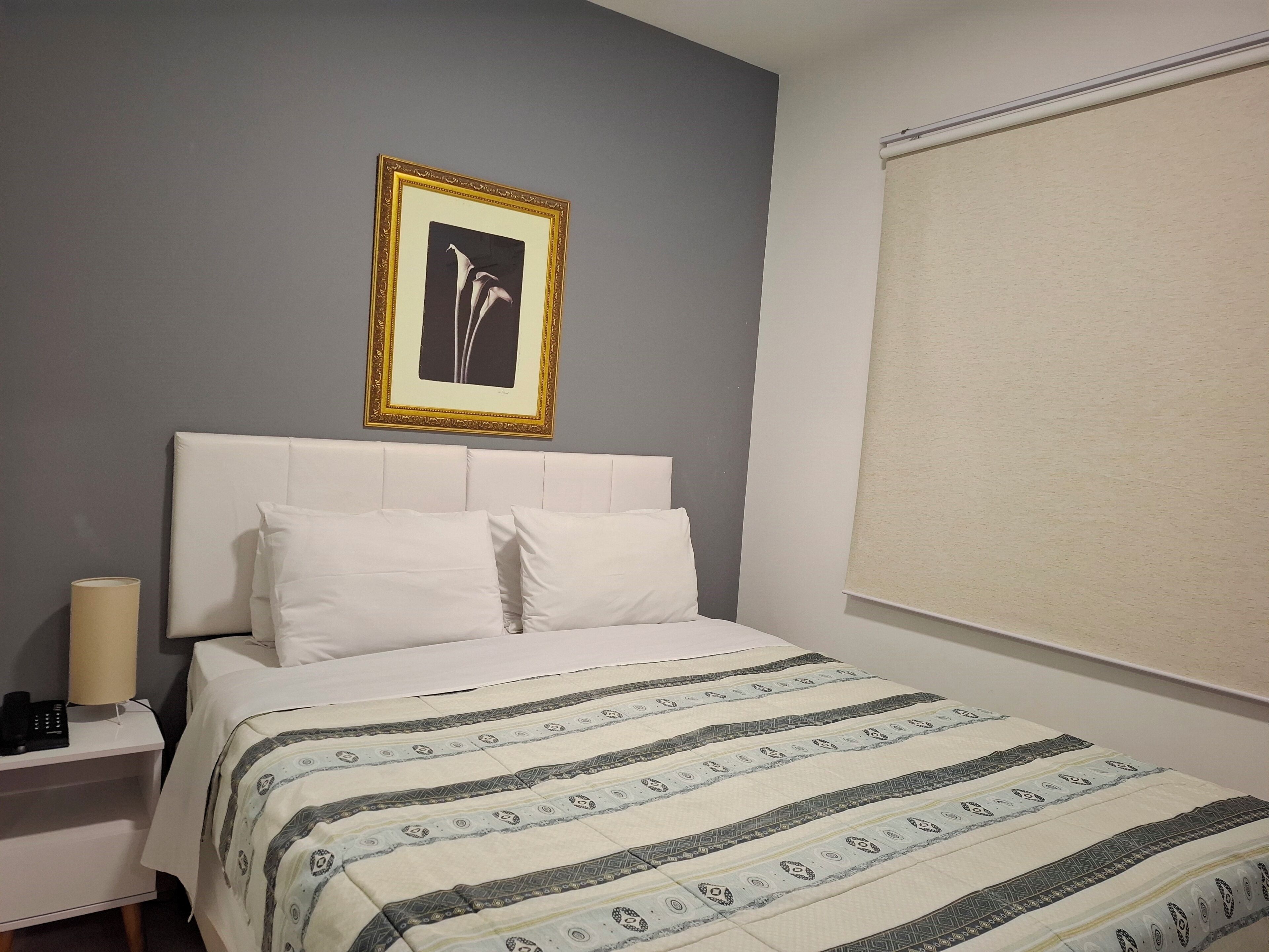 Quarto de hotel com cama, mesa de cabeceira, luminária, quadro emoldurado e janela com persiana.