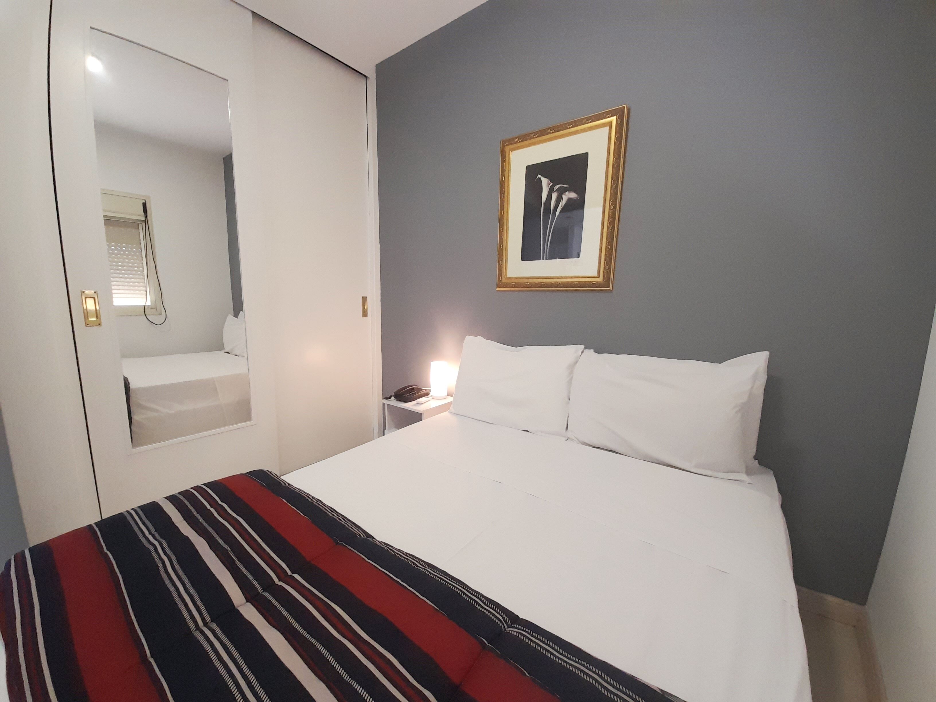 Ein Hotelzimmer mit Bett, Nachttisch mit Lampe, einem gerahmten Bild an der Wand und einer Tür mit Spiegel.