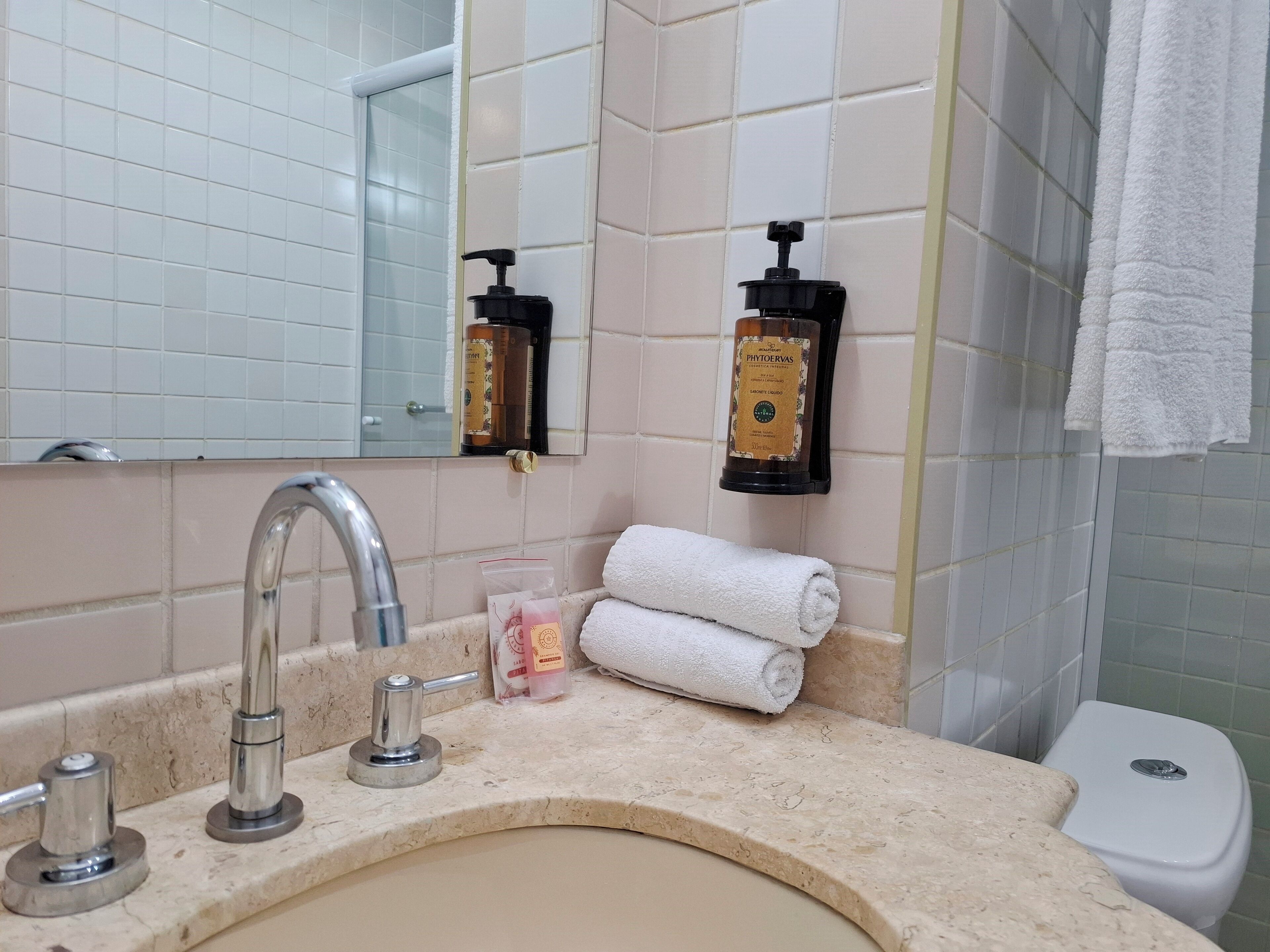 A bathroom with a sink, two faucets, a mirror, a towel rack with a rolled towel, and two soap dispensers.