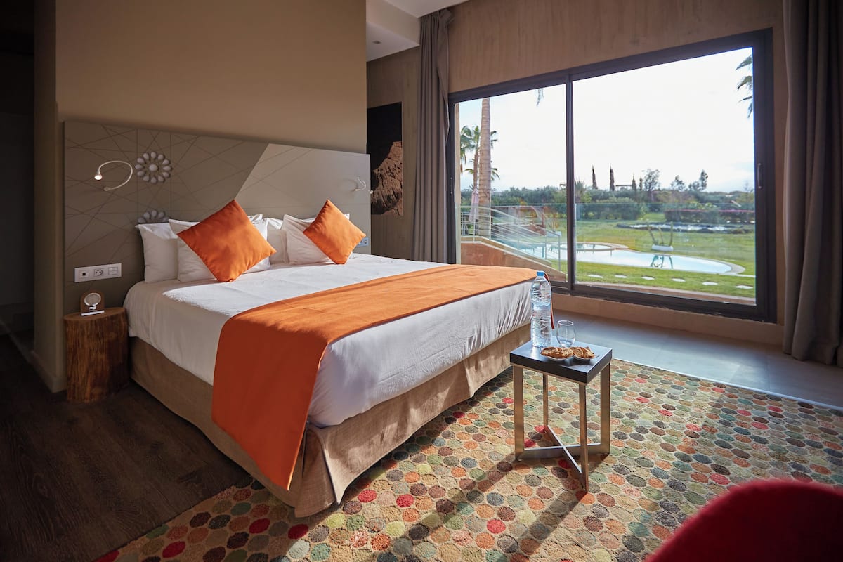 Habitación de hotel con una cama grande, una mesita con un vaso de agua y vistas a un área ajardinada a través de un ventanal amplio.