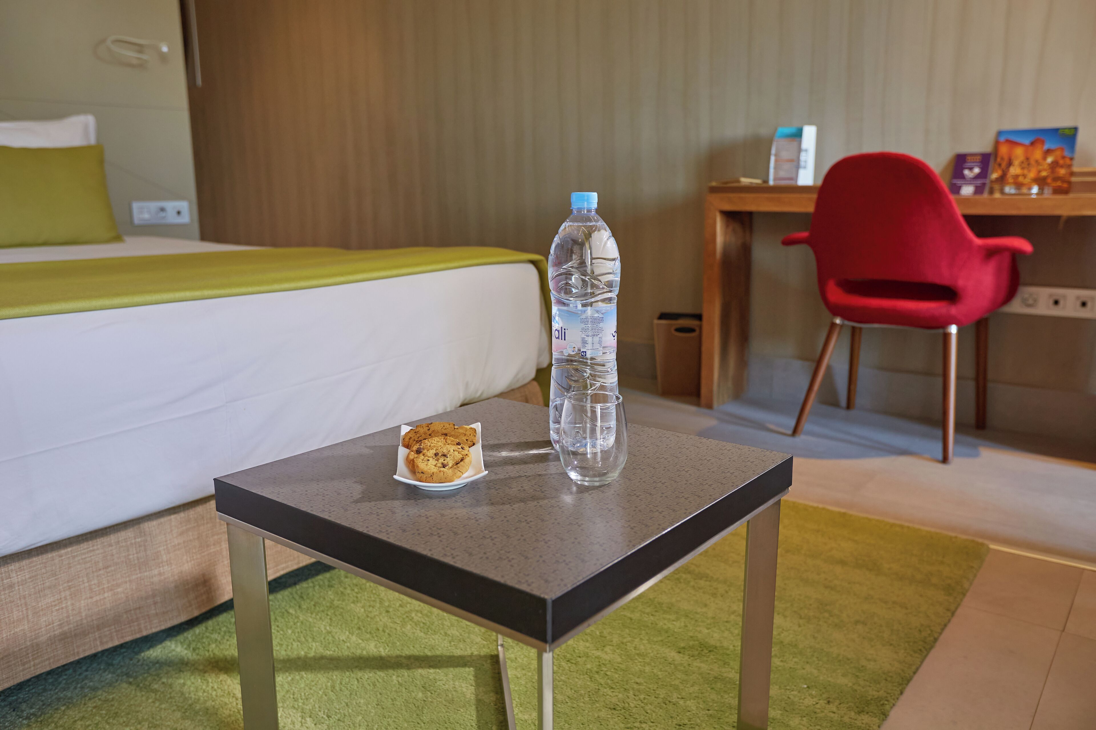 A hotel room with a bed, a small table with a bottle of water and cookies, a red chair, and a desk with a book and a lamp.
