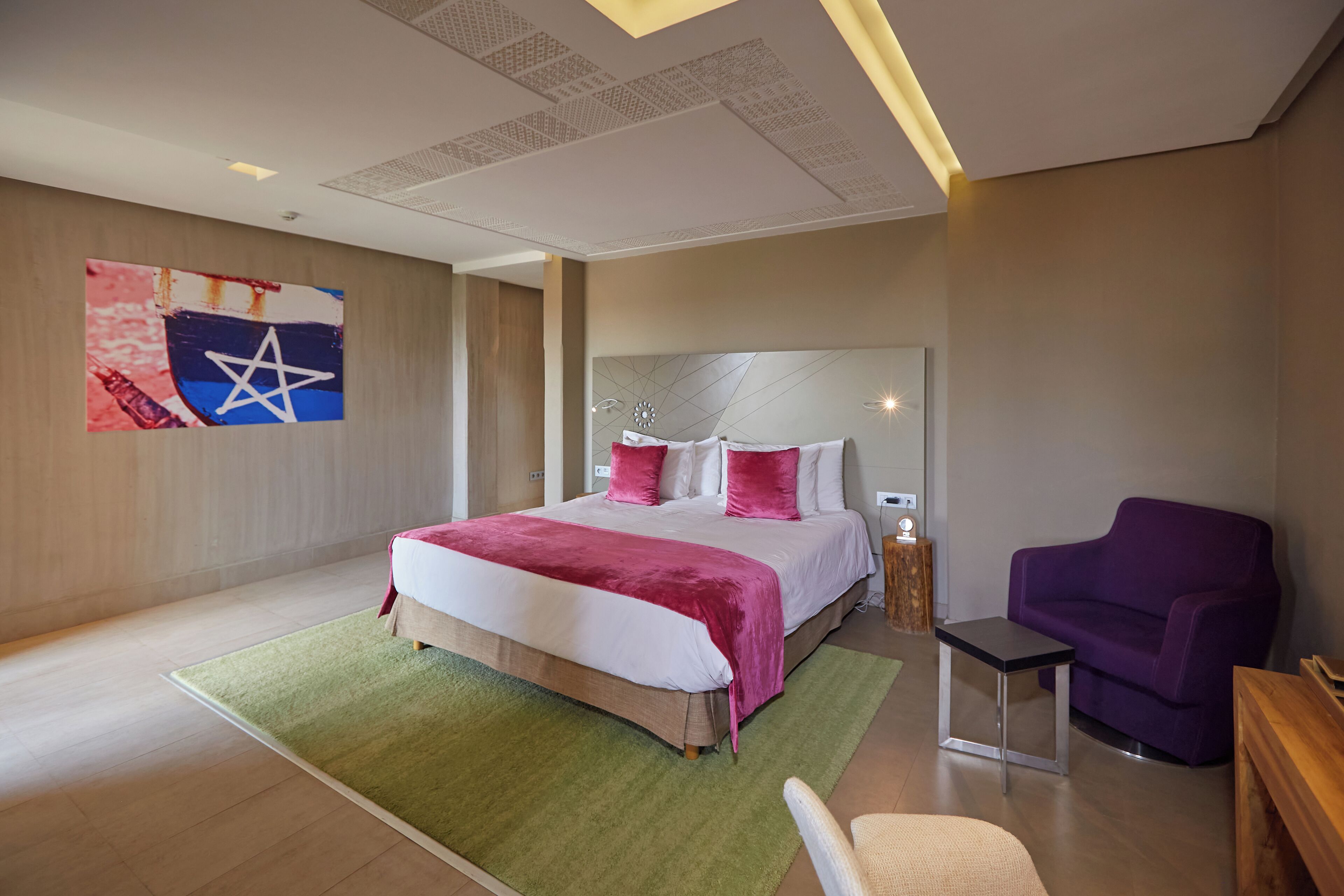 A modern hotel room with a large bed, a green rug, a purple armchair, and a small table.
