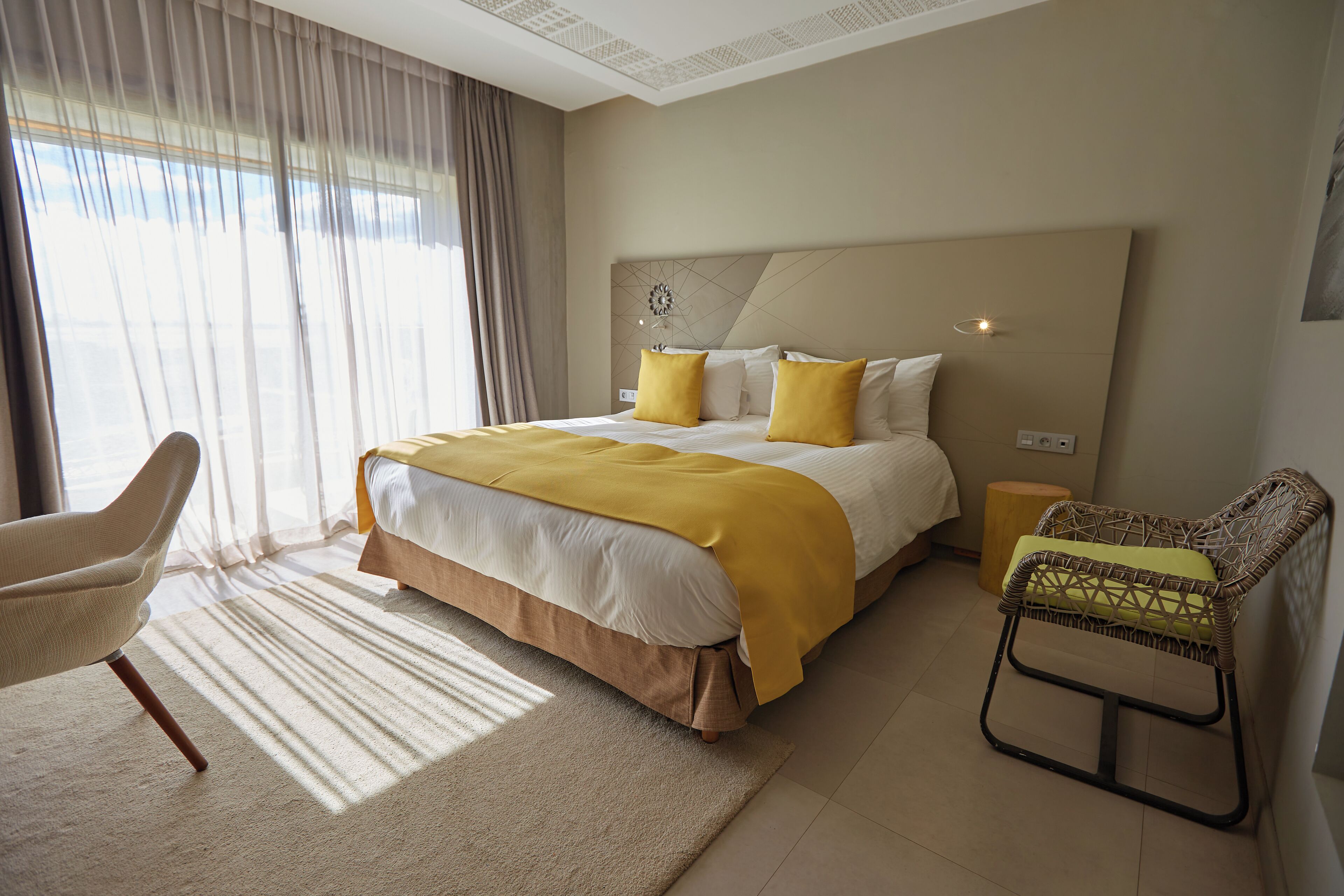 Habitación de hotel con una cama grande, una silla, una mesita de noche y un ventanal con cortinas.