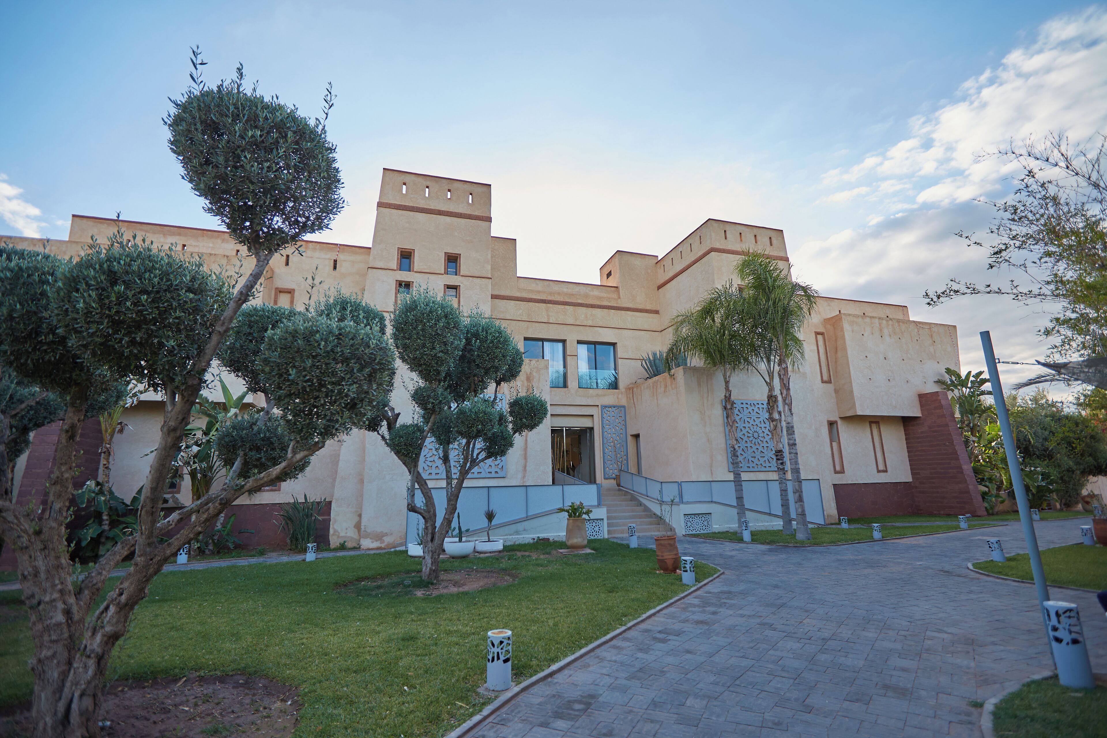A modern building with a symmetrical facade, a central entrance with steps, and a paved courtyard with neatly trimmed grass and trees.