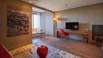 A modern hotel room with a large painting, a desk with a computer, a TV, and a red chair.