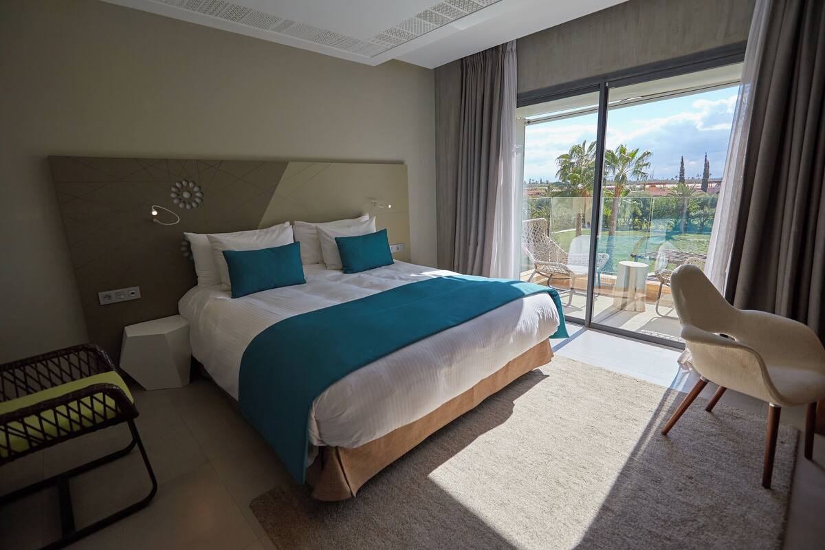 Habitación de hotel con una cama grande, una silla, una mesita y vistas a una piscina y palmeras.