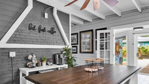 Une cuisine avec une table en bois foncé, des meubles blancs et un mur en briques décoratif orné d’une pancarte indiquant : « d’abord, un café ».