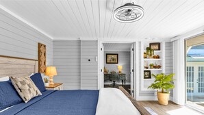 Une chambre avec un grand lit, une tête de lit en bois, une table de chevet avec une lampe, un bureau avec une chaise et une vue sur l’extérieur grâce à une porte vitrée.