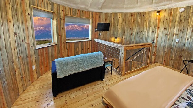 A room with wooden walls, a bed, a bench with a blanket, a fireplace, and a TV mounted on the wall. - Up-cycled Grain Bin Villa just 20 mins. from the Great Sand Dunes NP w/ WiFi, AC (Mosca)