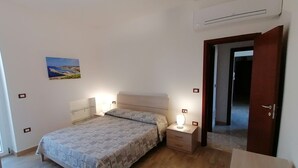 Un dormitorio con cama, mesitas de noche, lámpara de noche, aire acondicionado y una puerta que da a otra habitación.