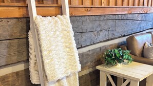 Interior em madeira com uma escada branca encostada à parede, um cobertor bege sobre ela, uma mesa de madeira com uma planta e um sofá com almofadas.