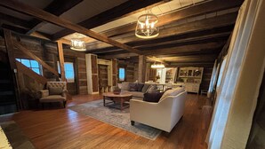Um amplo loft com vigas de madeira aparentes, um sofá branco grande, uma mesa de centro de madeira e um candelabro.