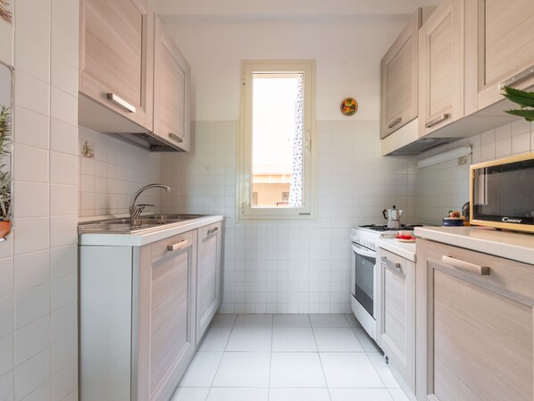A narrow kitchen with white tiles, wooden cabinets, a window, and a microwave.