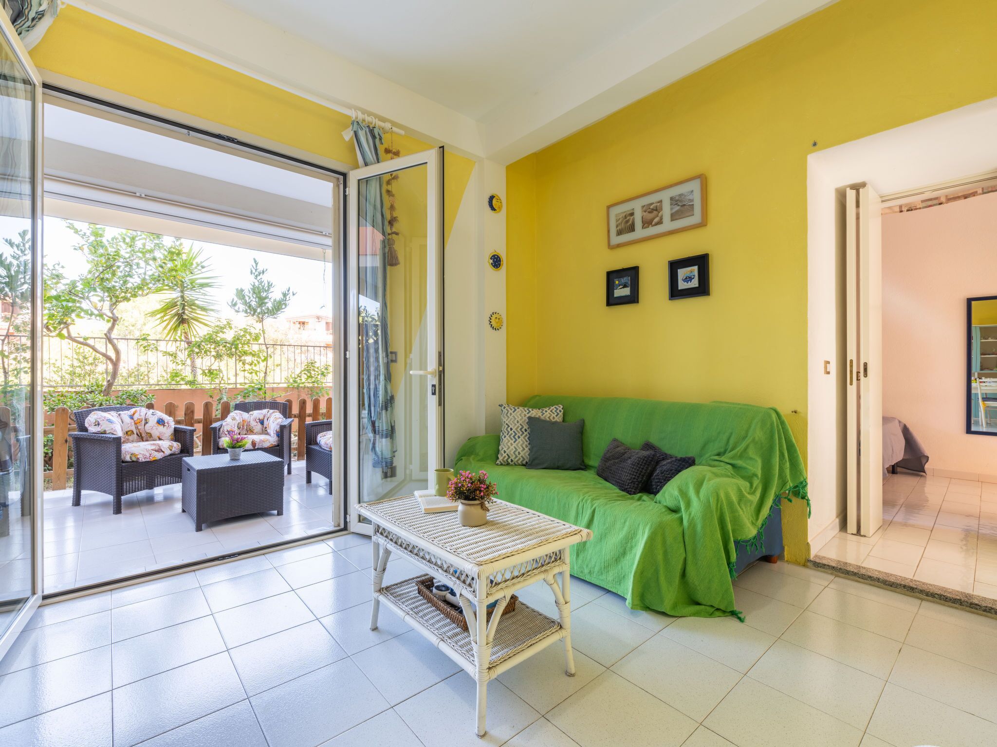 A bright living room with a green sofa, a wicker coffee table, and a glass door leading to an outdoor patio with wicker furniture and a view of trees.