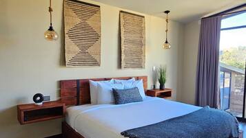 A modern bedroom with a large bed, wooden headboard, two bedside tables, a patterned rug, and a view of the outdoors through a window.