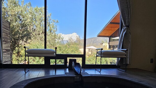 A swimming pool with two chairs and a table, large windows with curtains, and a view of trees and mountains. - CASA SEKUI (Valle de Guadalupe)