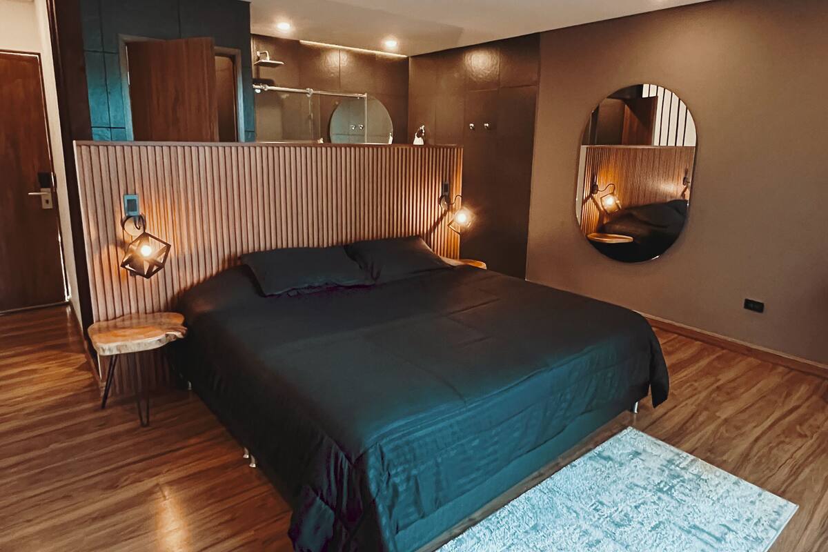 A modern bedroom with a large bed, wooden flooring, a round mirror, and a small table.
