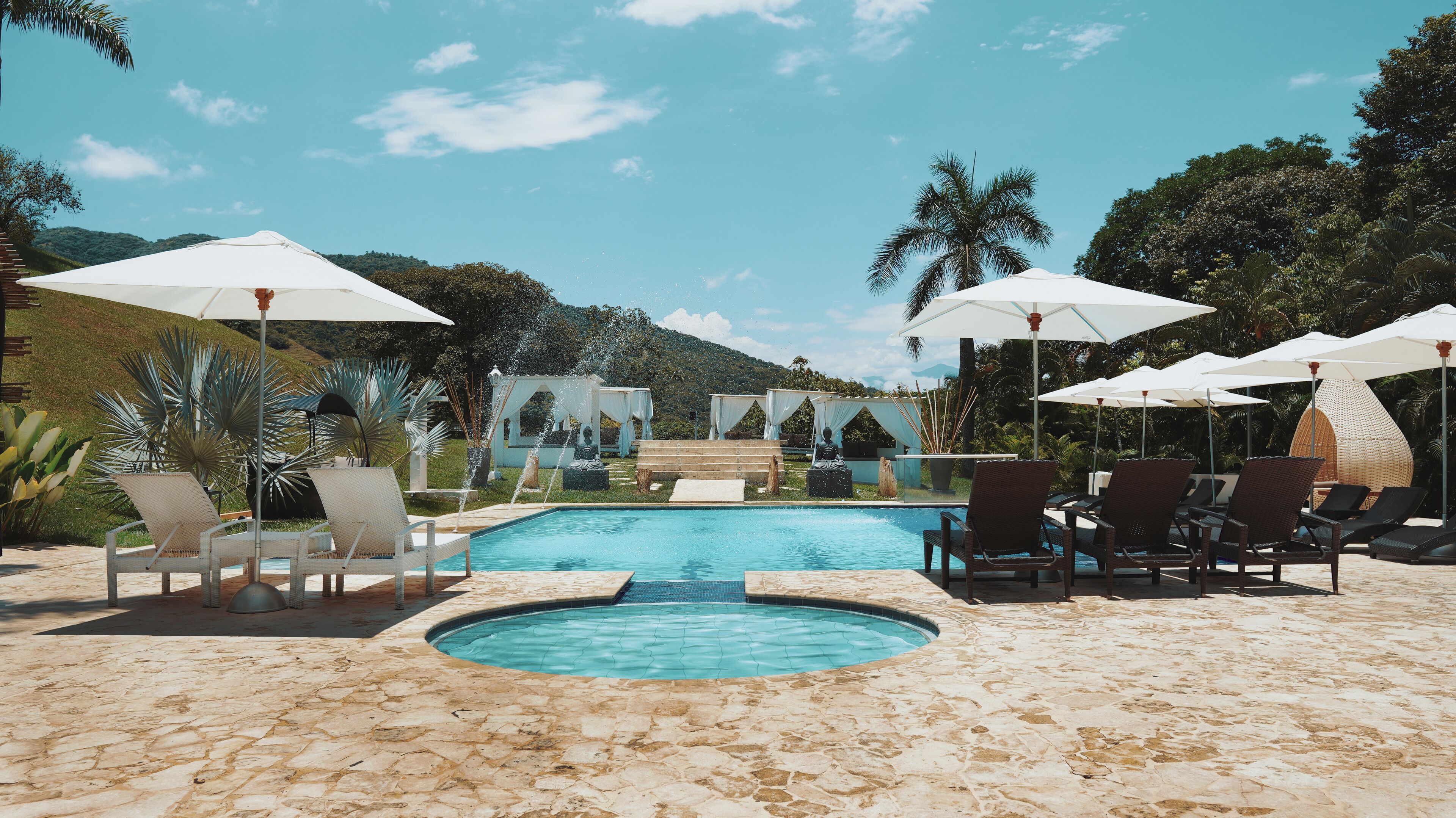 A pool area with lounge chairs and umbrellas, surrounded by palm trees and a mountainous backdrop.