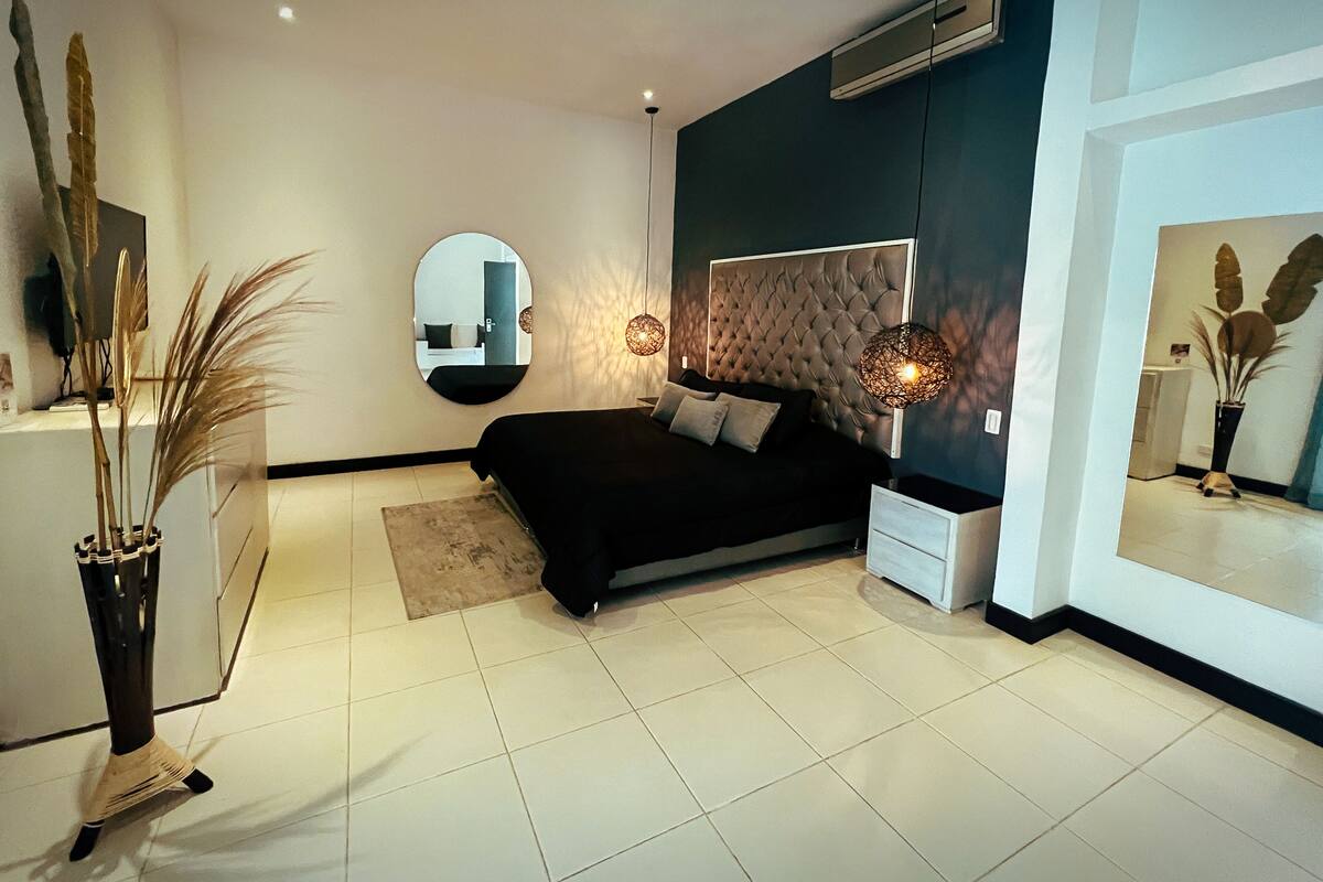 A modern bedroom with a large bed, bedside tables, a round mirror, and a potted plant.