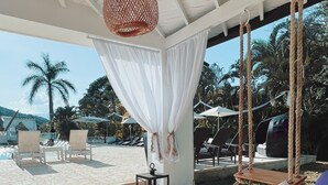 Een kamer aan het strand met een bed, een hanglamp en uitzicht op het strand en palmbomen.