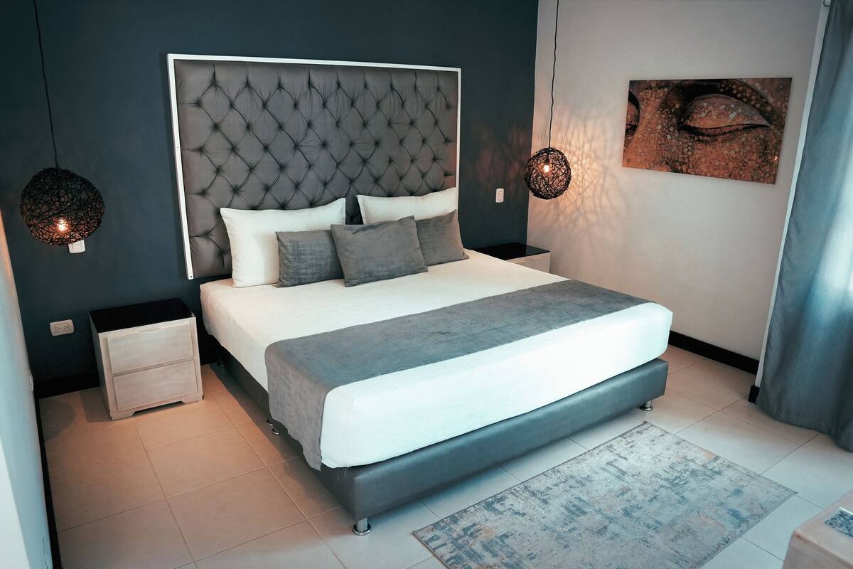 A modern bedroom with a tufted headboard, a large bed with grey bedding, two bedside tables, and a wall-mounted artwork.