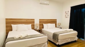 Una camera d'albergo con due letti, testiere in legno, biancheria bianca, un piccolo comodino con una lampada e un condizionatore d'aria a parete.