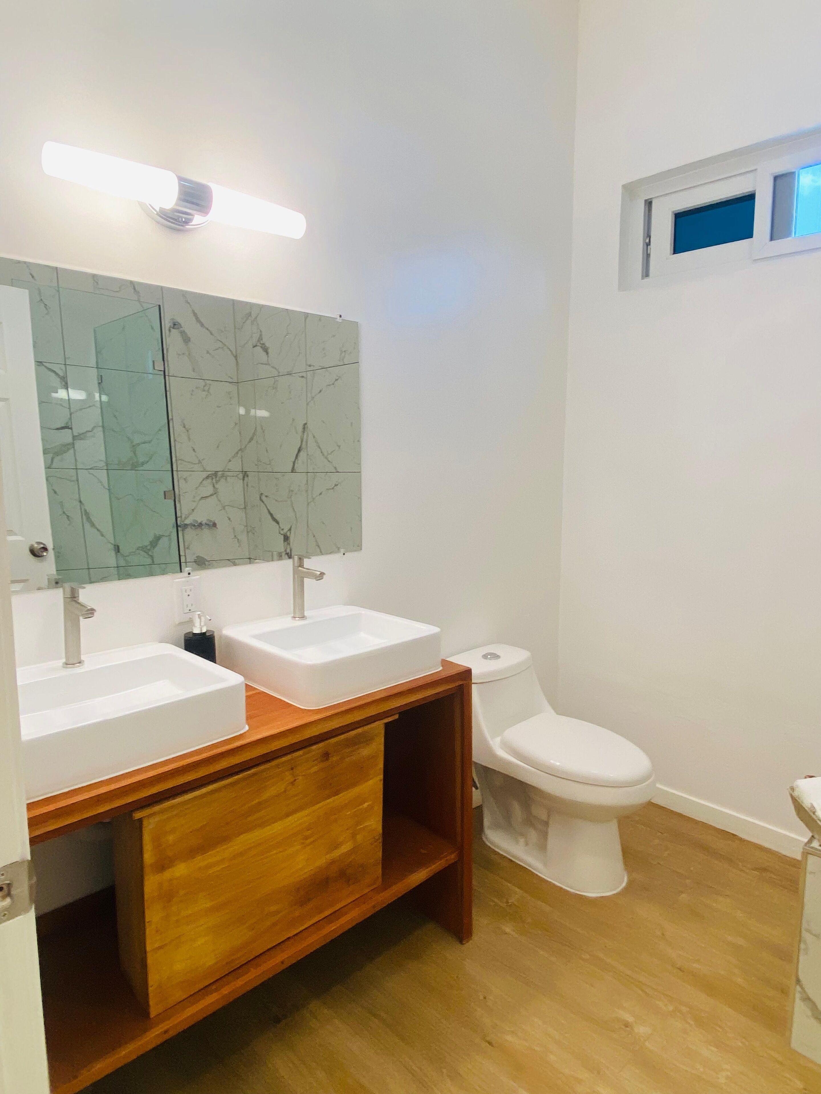A bathroom with two white sinks, a wooden vanity, a glass-tiled wall, and a white toilet.