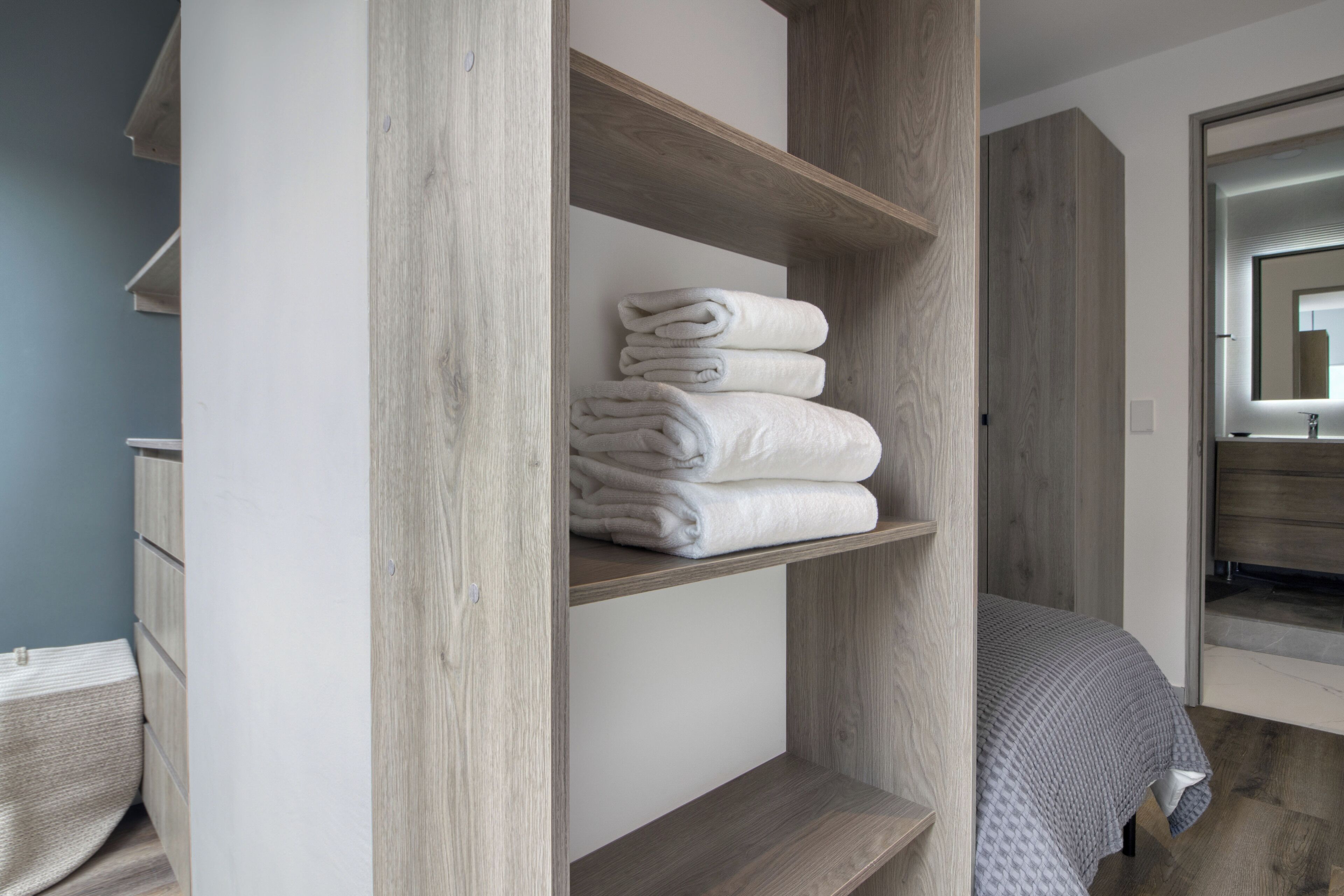 Un dormitorio moderno con un armario de madera, una cama con colcha estampada gris y un baño visible a través de una puerta abierta.