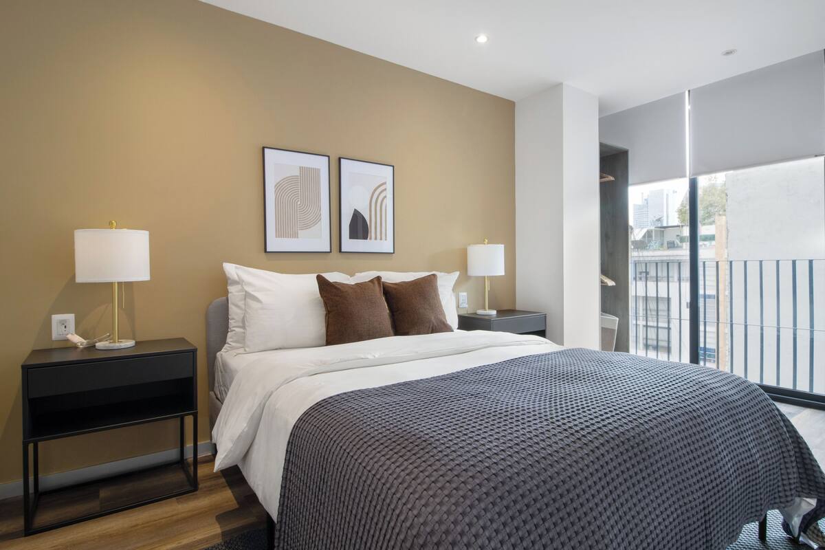 A modern hotel room with a large bed, two bedside tables with lamps, a view of the city through a balcony, and abstract wall art.