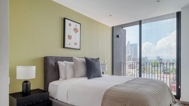 A modern hotel room with a large bed, a nightstand, a lamp, and a city view through the window. - Blueground Ava Rio Rhin in Mexico City (Mexico City)
