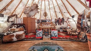 Un interior acogedor de una estructura circular tipo tienda de campaña, con una cama de madera en el centro, asientos cómodos y diversos elementos decorativos como alfombras y pieles de animales.