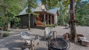 Una cabaña rústica con porche, rodeada de árboles. Se ven sillones Adirondack y una parrilla en primer plano.