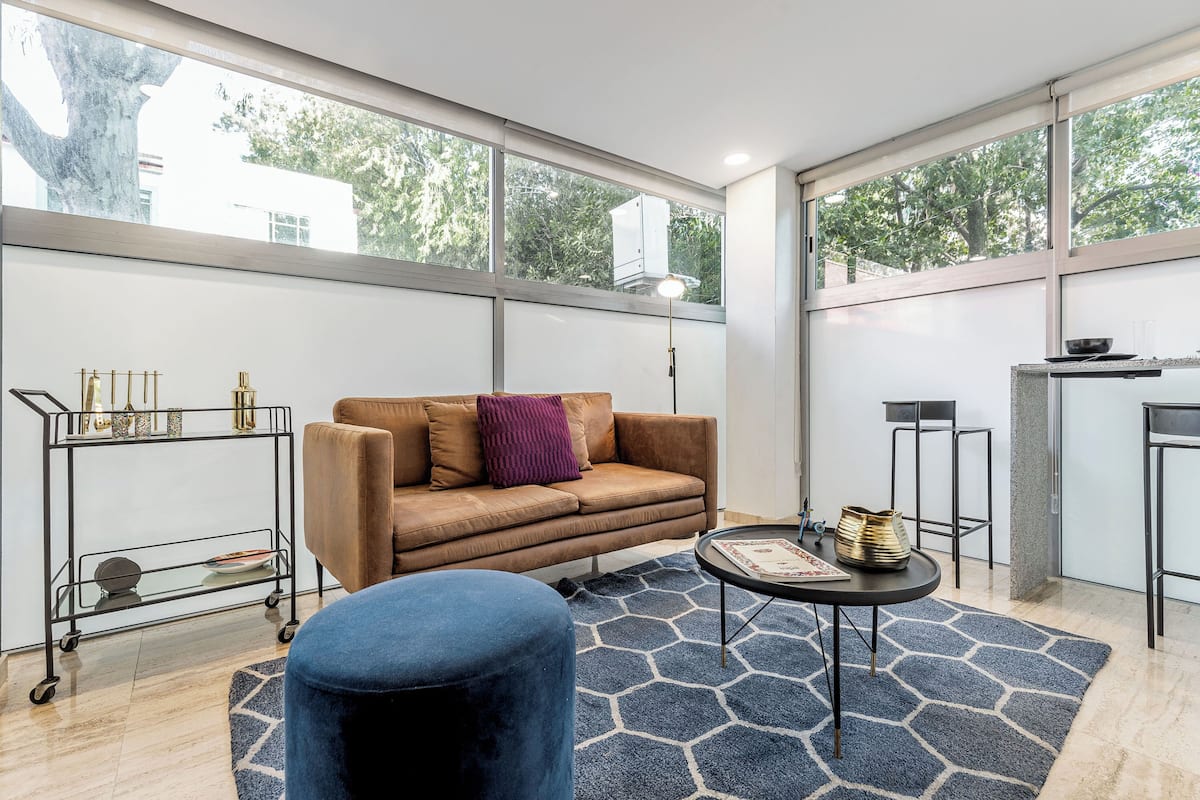 A modern living room with a sofa, ottoman, and a coffee table. There is a window providing natural light and a view of trees.