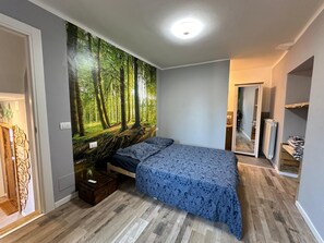 Makuuhuoneessa on suuri metsäaiheinen seinämaalaus, kuviollinen päiväpeite sängyllä, puinen yöpöytä ja patteri.