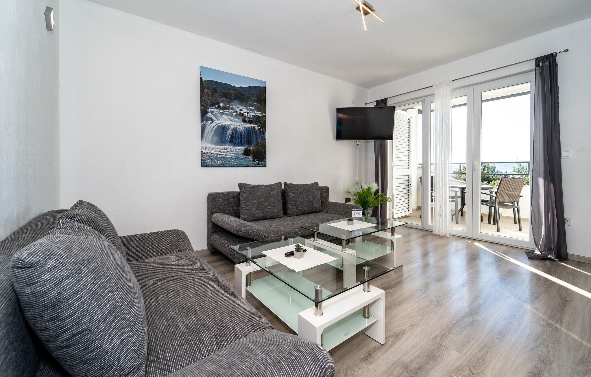 Een moderne woonkamer met een grijze bank, een glazen salontafel en een grote tv aan de muur.