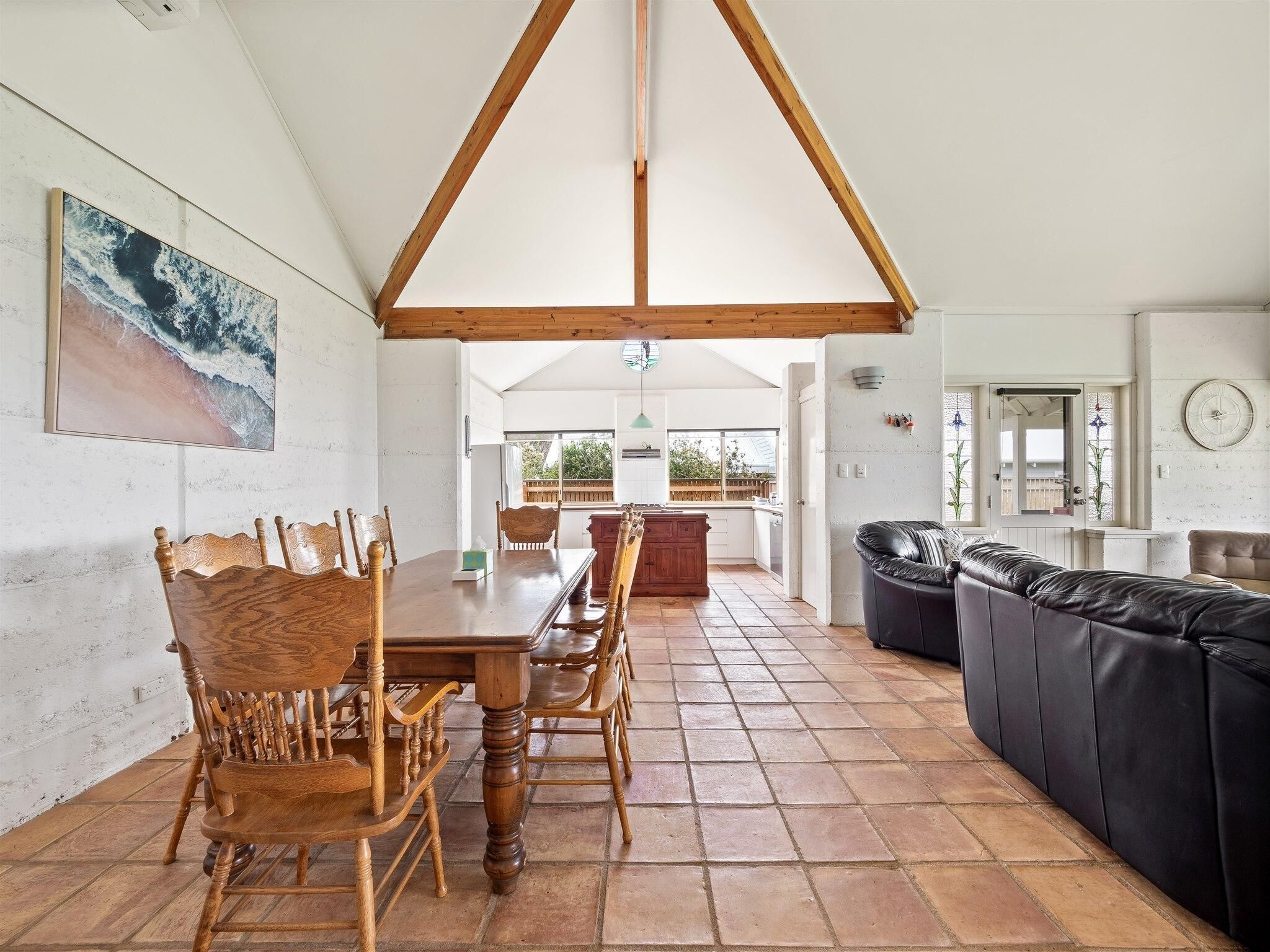 A spacious living area with a wooden dining table and chairs, a black leather sofa, and a kitchen in the background.