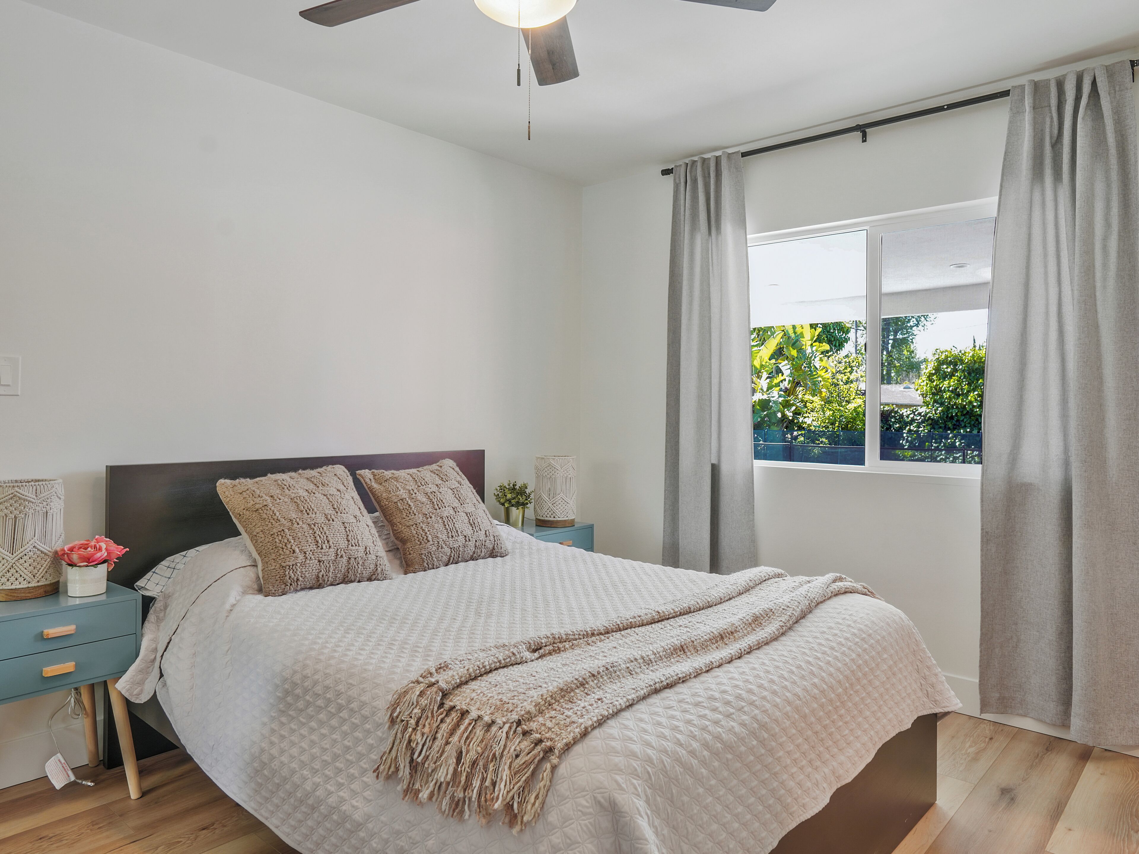 A bedroom with a bed, nightstands, a ceiling fan, a window with curtains, and a view of greenery outside.