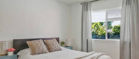 A bedroom with a bed, nightstands, a ceiling fan, a window with curtains, and a view of greenery outside.