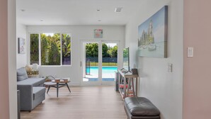 Un vaste espace de vie comprenant un canapé gris, une table basse et un banc. On aperçoit une piscine à travers les portes vitrées, et des tableaux encadrés ornent les murs.