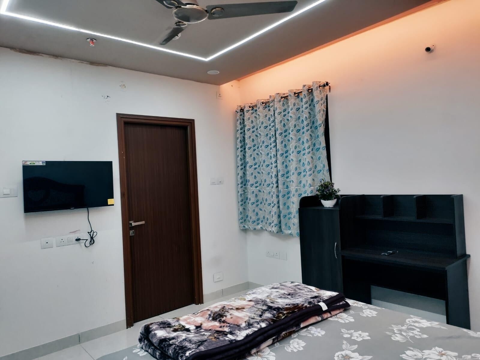 A bedroom with a bed, a TV mounted on the wall, a ceiling fan, and a door.