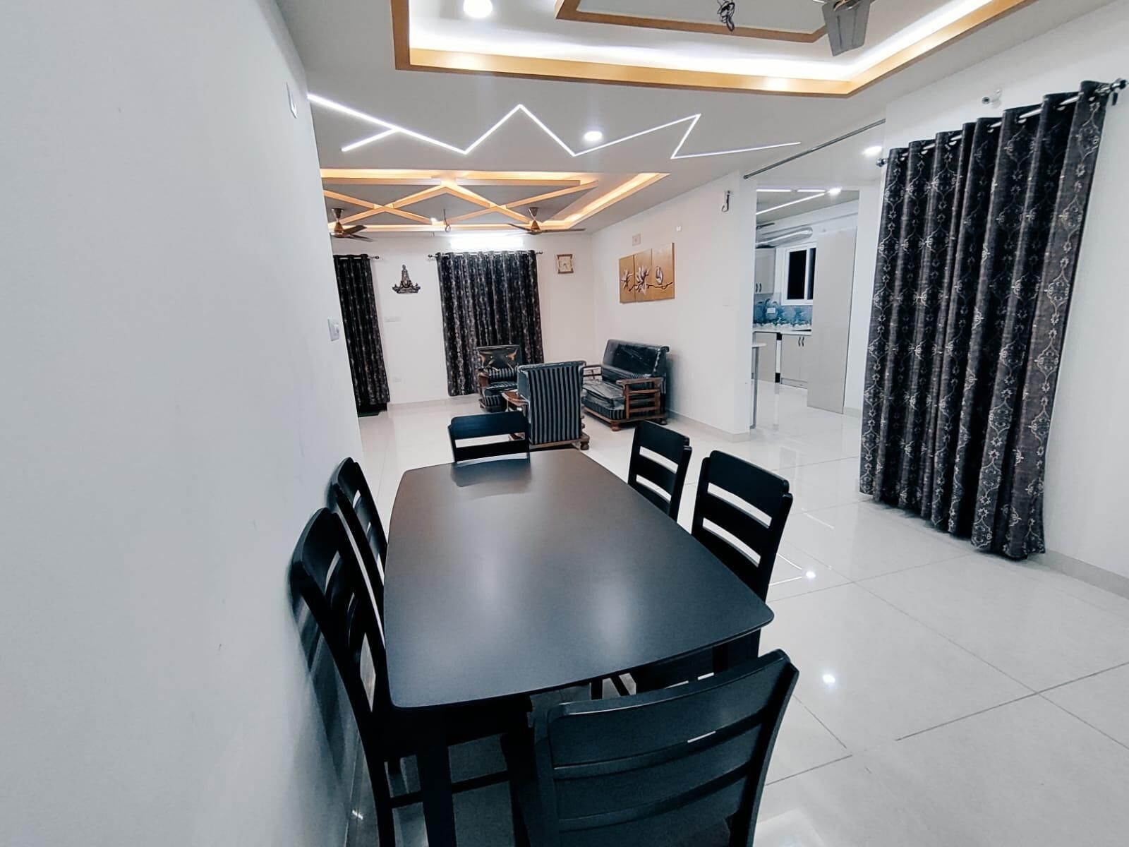 A modern dining area with a rectangular table and black chairs, a ceiling with geometric lighting, and a kitchen in the background.