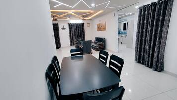 A modern dining area with a rectangular table and black chairs, a ceiling with geometric lighting, and a kitchen in the background.