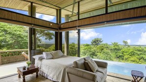 A modern bedroom with a large bed, a sofa, and a view of lush greenery. - Villa La Loma, Oceanview, Private Pool and AC! (Tarcoles)