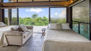 A modern bedroom with two beds, a sofa, and a small table. Large windows offer a view of greenery. - Villa La Loma, Oceanview, Private Pool and AC! (Tarcoles)