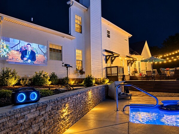 A well-lit outdoor area with a pool, a stone wall, and a large screen displaying a news broadcast.