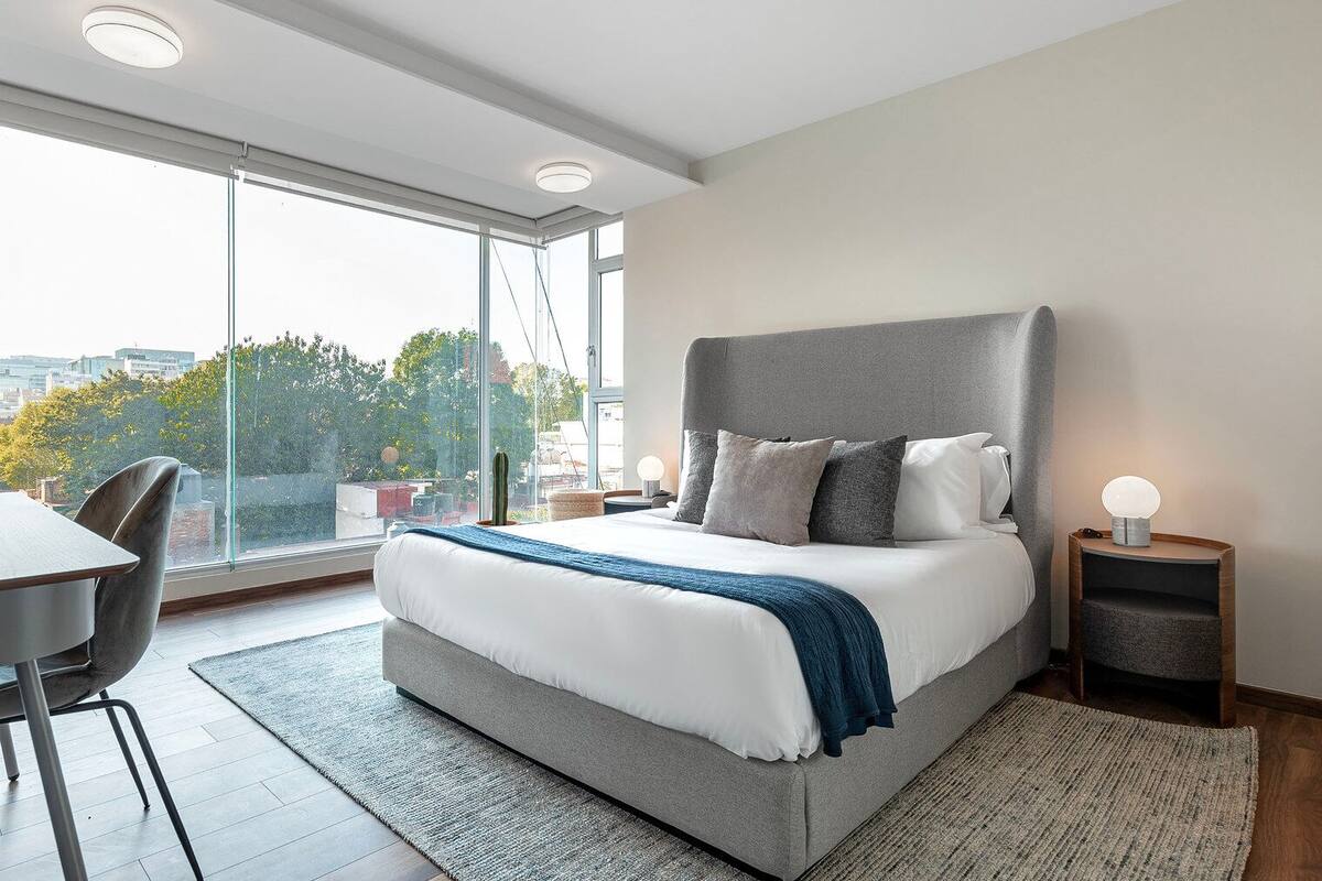 A modern bedroom with a large bed, a desk, and a view of the city through large windows.